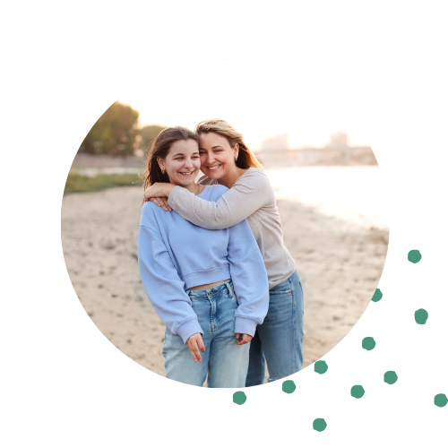 Mom hugging teen daughter on the beach. The teen is wearing a light blue sweatshirt and the mom is wearing a tan shirt. Both are wearing jeans and have blonde hair.