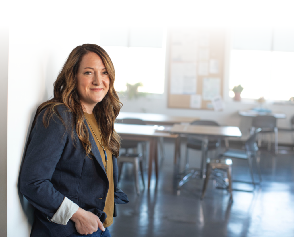 Woman stands in front of classroom door with desks and chairs in background. The woman has curly brown hair, a blue blazer, and a burnt orange top on.